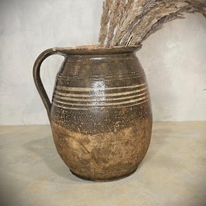 Può includere: Una brocca rustica in ceramica marrone con manico, caratterizzata da strisce bianche orizzontali. La brocca è riempita con erba ornamentale beige essiccata, creando un'estetica naturale e terrosa. La brocca ha un aspetto invecchiato.
