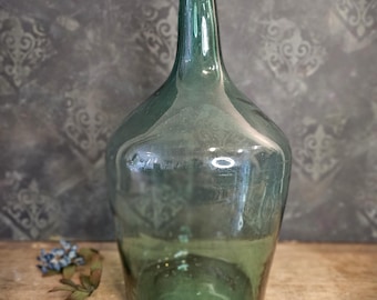 Puede incluir: Una gran botella de vidrio verde transparente con una boca ancha y un fondo redondeado. La botella está vacía y tiene un ligero tono verde.