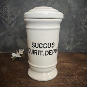 Może przedstawiać: Biały ceramiczny słoik z pokrywką i czarnym napisem "SUCCUS QUIRIT. DEPUR."