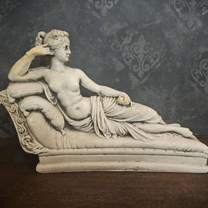 May include: A white plaster sculpture of a reclining woman in a draped gown, resting on a chaise lounge with ornate details.