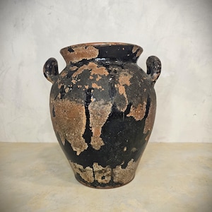 May include: A vintage, dark-colored ceramic vase with two side handles. The vase has a black base with brown and tan distressed paint. The rim is a lighter brown color. The vase is likely used for decorative purposes.