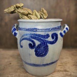 May include: A white ceramic crock with blue and white painted designs. The crock has two handles and is decorated with a blue swirl pattern.