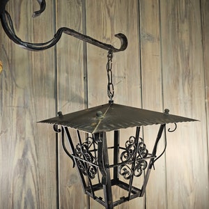 May include: A black metal lantern with a decorative design hangs from a chain attached to a wrought iron hook. The lantern has a square shape with a pointed top and a decorative design on the sides.