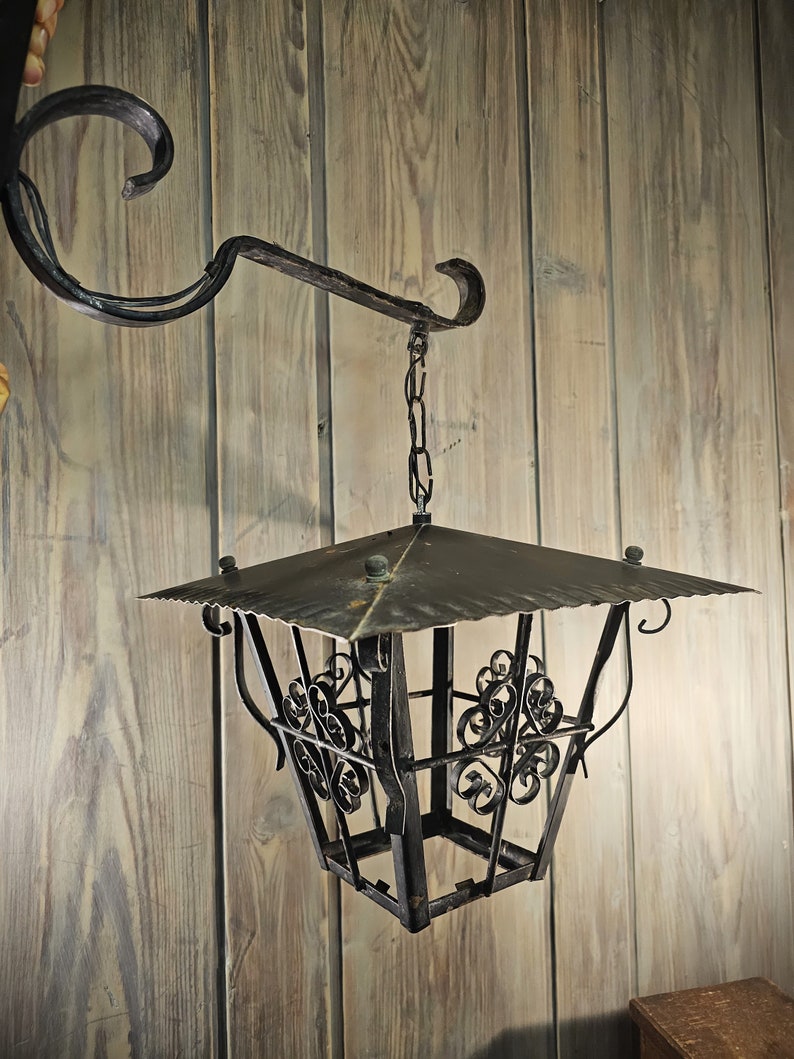 May include: A black metal lantern with a decorative design hangs from a chain attached to a wrought iron hook. The lantern has a square shape with a pointed top and a decorative design on the sides.