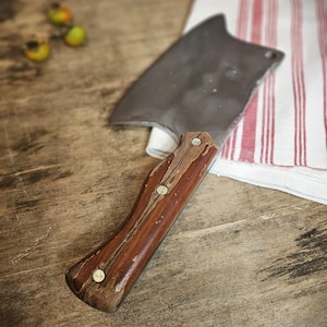 Op de afbeelding: Een vintage hakmes met een houten handvat en een metalen blad. Het hakmes ligt op een wit en rood gestreepte keukendoek.