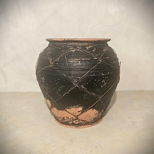 Puede incluir: Una olla redonda de cerámica negra con una jaula de alambre con diseño de diamantes. La olla tiene una boca ancha y una superficie texturizada. La base y el borde son de color marrón claro. La olla mide aproximadamente 25 cm de altura.