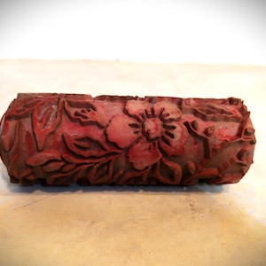 May include: A cylindrical paint roller sleeve featuring a raised, embossed floral pattern with detailed petals and leaves. The roller is coloured in a deep, muted red tone and has a textured surface designed for applying decorative patterns to surfaces.