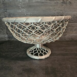 May include: A silver wire mesh bowl with a pedestal base. The bowl is designed with a woven wire pattern and has a rounded shape.