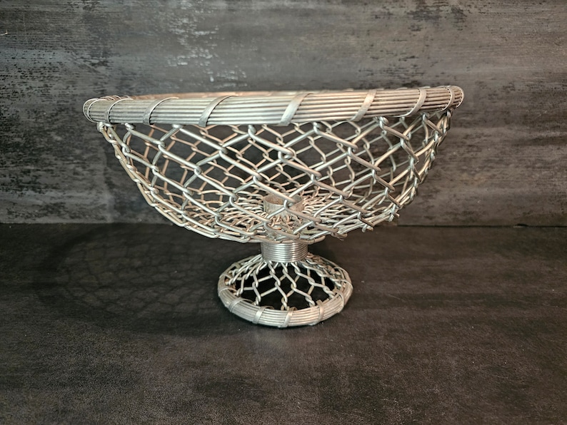 May include: A silver wire mesh bowl with a pedestal base. The bowl is designed with a woven wire pattern and has a rounded shape.