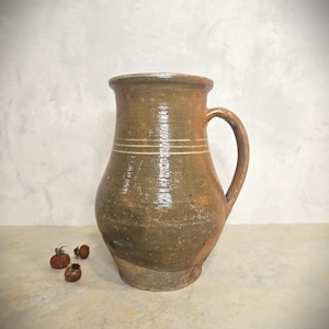 May include: A rustic, brown ceramic pitcher with a handle. The pitcher has a wide base, a rounded body, and a narrow neck. Two horizontal white stripes encircle the body. Small, dried seed pods are in the foreground.