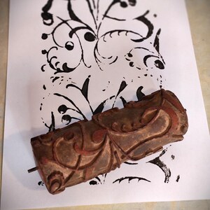 Pode incluir: Um rolo de pintura cilíndrico com uma superfície castanha desgastada e detalhes em relevo. O rolo está posicionado sobre uma folha de papel branca com uma transferência de tinta preta do mesmo design floral complexo e ondulado.