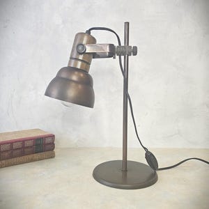 May include: A vintage desk lamp in a bronze finish. The lamp has an adjustable head, a circular base, and a vertical support. A black power cable extends from the base. The lamp is on a light surface, with books in the background. The lamp's height is approximately 30 cm.