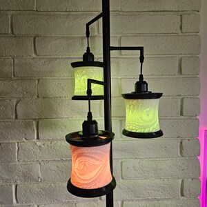Può includere: Una lampada da terra nera con tre paralumi. Ogni paralume è cilindrico con un design intagliato dettagliato ed emette una luce soffusa colorata. La lampada è posizionata davanti a un muro di mattoni bianchi.
