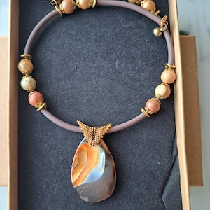 May include: A brown leather necklace with a large, teardrop-shaped pendant made of agate. The pendant has brown, orange, and white swirls. The necklace is adorned with small, round beads in shades of brown, beige, and pink.