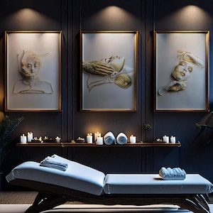 Op de afbeelding: Een spa-interieur met drie ingelijste kunstwerken met gouden accenten, die gestileerde gezichten tonen. Een massagetafel met een wit kussen staat voor de kunstwerken. Kaarsen en opgerolde handdoeken staan op een plank, wat een ontspannen sfeer creëert.