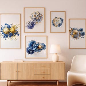 May include: Five framed watercolour art prints showcasing abstract botanical designs in blue, gold, and white. The prints are displayed above a light wood cabinet, a lamp, and a white chair.