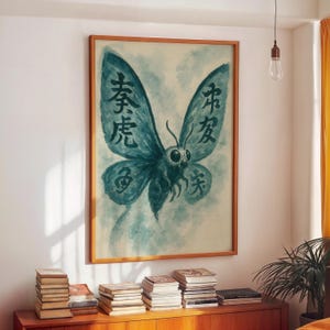 Op de afbeelding: Een ingelijst kunstwerk met een turquoise vlinder met Chinese karakters op zijn vleugels. De vlinder heeft grote ogen en staat tegen een lichtblauwe en witte achtergrond. De lijst heeft een warme bruine kleur.