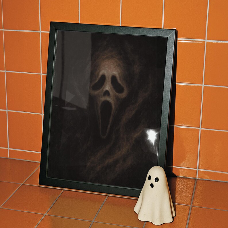 May include: A framed print featuring the Scream mask from the horror film series, with a small ghost figurine in front. The print is in a black frame, and the ghost figurine is white with black eyes. The background is orange.