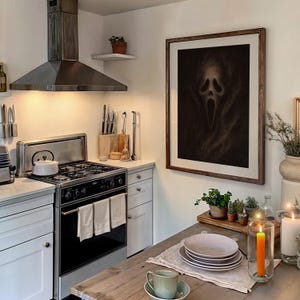 May include: A kitchen scene featuring a framed artwork of a screaming face. The kitchen has white cabinets, a stainless steel range hood, and a wooden table set with plates and a teacup. Candles and plants add to the decor.