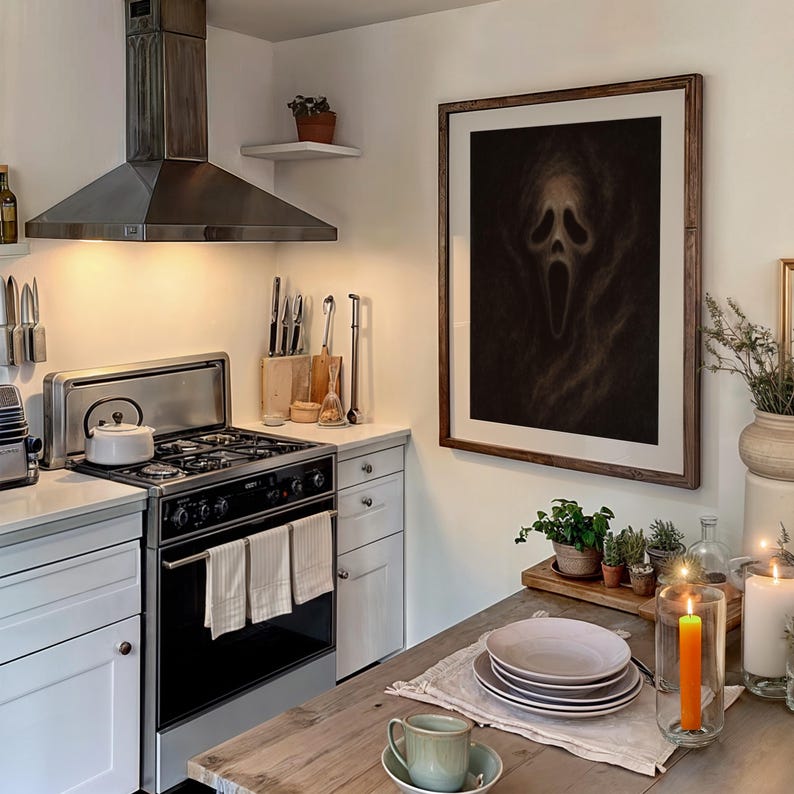 May include: A kitchen scene featuring a framed artwork of a screaming face. The kitchen has white cabinets, a stainless steel range hood, and a wooden table set with plates and a teacup. Candles and plants add to the decor.