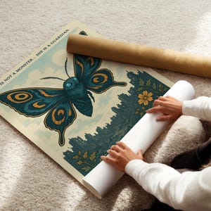 May include: A poster featuring a large teal and gold butterfly illustration with the text "SHE IS NOT A MONSTER... SHE IS A GUARDIAN." The poster is rolled up with a brown tube and a white tube.