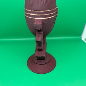 May include: A dark maroon, egg-shaped object with a textured, vertical-lined surface. It features three horizontal carved grooves near the top and a series of three protruding, block-like rectangular elements extending from the side, resting on a circular base.