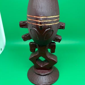 May include: A dark brown, carved wooden decorative vase with a textured surface. The vase has a rounded top with horizontal gold bands and a stylized face design. The base is round and the overall design is reminiscent of tribal art.
