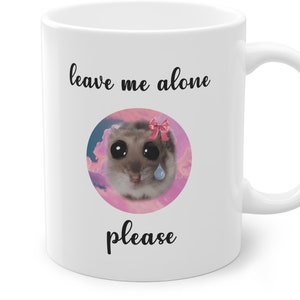 Può includere: Tazza in ceramica bianca con un'illustrazione in bianco e nero di un criceto con un fiocco rosa e una singola lacrima. Il testo "leave me alone please" è stampato sulla tazza.