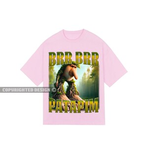 May include: Pink t-shirt featuring a cartoon creature with a long nose and green vines. The text "BRR BRR PATAPIM" is printed in gold on the shirt.