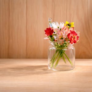 May include: A small glass jar filled with colourful glass flowers. The flowers include red, white, pink, and yellow blooms. The jar sits on a wooden surface with a wooden background.