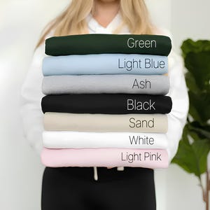 Puede incluir: Una pila de ocho camisetas de diferentes colores, incluyendo verde, azul claro, ceniza, negro, arena, blanco y rosa claro. Cada camiseta tiene el nombre del color escrito en ella.