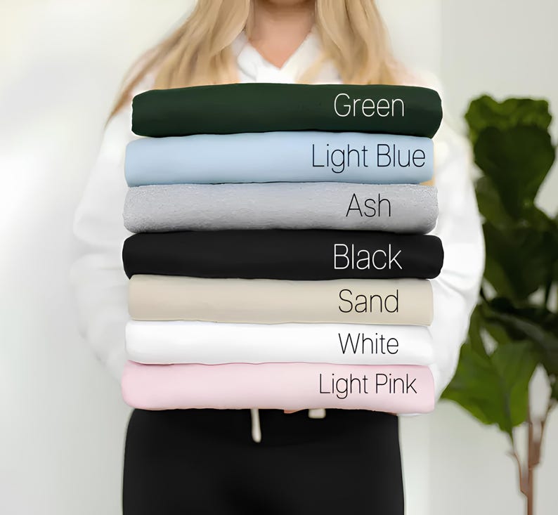Puede incluir: Una pila de ocho camisetas de diferentes colores, incluyendo verde, azul claro, ceniza, negro, arena, blanco y rosa claro. Cada camiseta tiene el nombre del color escrito en ella.