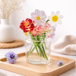 May include: A small glass jar filled with colourful glass flowers, including red, pink, and white daisies, sits on a wooden tray. A single blue flower and a white vase with dried flowers are also present.
