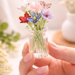 May include: A small glass vase containing a collection of colourful glass flowers. The flowers are in shades of red, pink, blue, purple, and white. The clear vase has a ribbed design. The flowers are held in a hand.