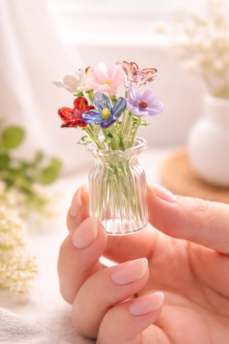 May include: A small glass vase containing a collection of colourful glass flowers. The flowers are in shades of red, pink, blue, purple, and white. The clear vase has a ribbed design. The flowers are held in a hand.