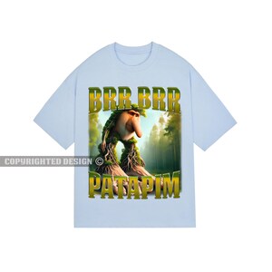 May include: A light blue t-shirt featuring a cartoon creature with a long nose and green hair, standing in a forest. The text "BRR BRR PATAPIM" is printed in yellow and green lettering on the shirt.