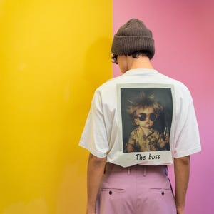 Puede incluir: Una camiseta blanca con un gr&aacute;fico de un ni&ntilde;o con gafas de sol y sosteniendo un vaso. El texto "The boss" est&aacute; impreso debajo del gr&aacute;fico. La persona lleva una boina marr&oacute;n y pantalones rosas.