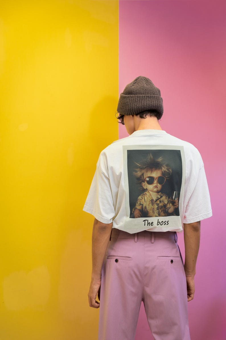 Puede incluir: Una camiseta blanca con un gr&aacute;fico de un ni&ntilde;o con gafas de sol y sosteniendo un vaso. El texto "The boss" est&aacute; impreso debajo del gr&aacute;fico. La persona lleva una boina marr&oacute;n y pantalones rosas.