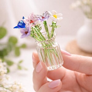 May include: A small glass vase containing glass flowers in shades of blue, purple, pink, and white. The flowers have green stems and are held in a hand. The vase is a miniature piece of glass art, showcasing intricate details.