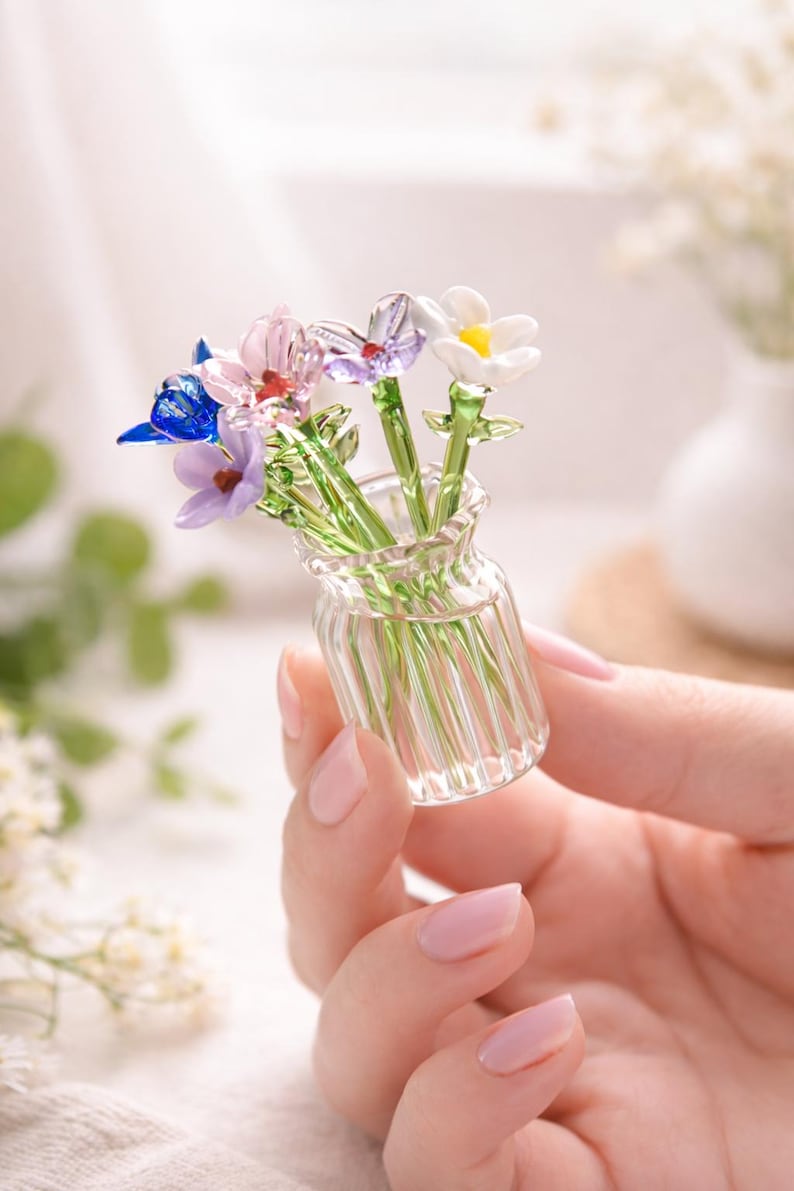May include: A small glass vase containing glass flowers in shades of blue, purple, pink, and white. The flowers have green stems and are held in a hand. The vase is a miniature piece of glass art, showcasing intricate details.