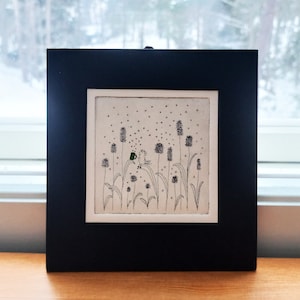 May include: A black framed artwork with a white background and a black and white illustration of a small person standing in a field of tall grass with small green flowers. The person is holding a green cup.