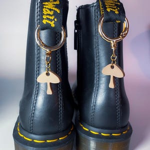 May include: Black leather boots with yellow stitching and a yellow and black pull tab. Each boot has a gold-colored charm with a mushroom pendant attached. The charms are hanging from a gold-colored ring.