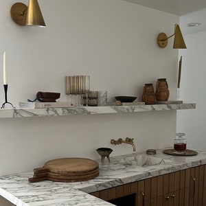 May include: A white marble countertop with a wooden cutting board and a gold faucet. The countertop is part of a kitchen island with wooden cabinets and a white marble backsplash. There are two gold wall sconces on the wall behind the countertop.