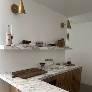 Puede incluir: Una encimera de mármol blanco con una tabla de cortar de madera y un fregadero. La encimera forma parte de una barra o isla de cocina con armarios de madera y un grifo dorado. Hay dos apliques de pared dorados con pantallas en forma de cono.