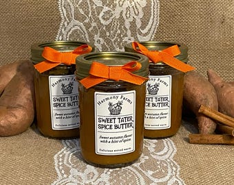 May include: Three glass jars of sweet potato spice butter, each with an orange bow. The labels read "Sweet Tater Spice Butter" and "Sweet autumn flavour with a hint of spice." The jars are on a lace cloth.