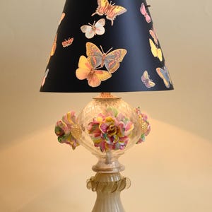 Può includere: Una lampada da tavolo con un paralume nero decorato con varie illustrazioni di farfalle colorate. La base è in vetro trasparente con accenti floreali tridimensionali multicolori e un supporto in vetro scanalato color bianco sporco.