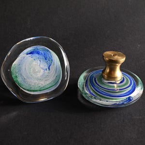 May include: Two circular glass cabinet knobs featuring swirling patterns of blue, green, and white. One knob is shown from the front, while the other is shown from the side, revealing a metallic gold-toned base with a central screw hole for mounting.