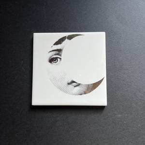 Può includere: Piastrella quadrata in ceramica bianca con un'illustrazione nera e argento di un volto parzialmente oscurato da una luna crescente. Il disegno comprende un occhio e labbra, con la forma della luna in argento. La piastrella è su una superficie nera.