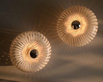 May include: Two circular wall lamps featuring a clear and white striped glass pattern radiating from a central polished chrome fixture. The lights emit a warm glow, casting soft, elongated shadows across the surrounding surface area.
