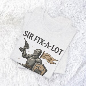 Peut inclure: T-shirt gris clair avec le texte "SIR FIX-A-LOT" au-dessus d'une illustration d'un chevalier tenant une perceuse. Le chevalier a un bouclier avec des outils et une banni&egrave;re qui dit "DUCTUS MAXIMUS".